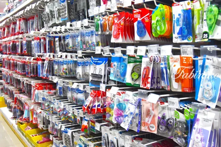 Aisle of a Malaysian stationery store displaying colorful correction tapes, paper clips, and other office supplies on white racks.