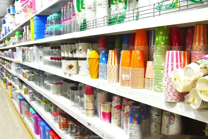 Colorful paper cups and plastic containers neatly arranged on white shelves in a Malaysian household goods store.