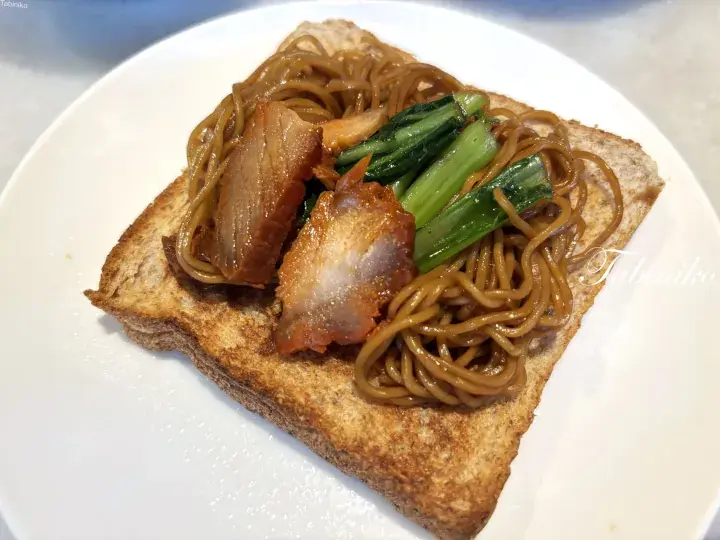 Crispy toast topped with savory noodles, roasted pork, and green vegetables on a white plate.