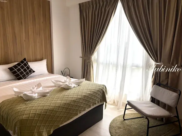 Cozy hotel bedroom with a neatly made bed featuring folded towels, wooden headboard, and sheer curtains.