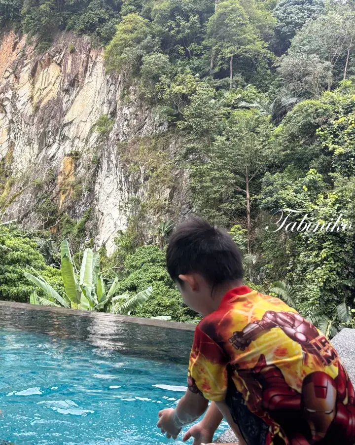 Child in Iron Man swimwear by an infinity pool overlooking a lush tropical cliffside.