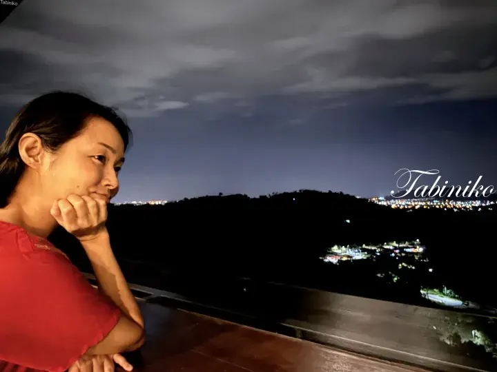 Woman enjoying a night view from a balcony overlooking a hillside town with scattered lights.
