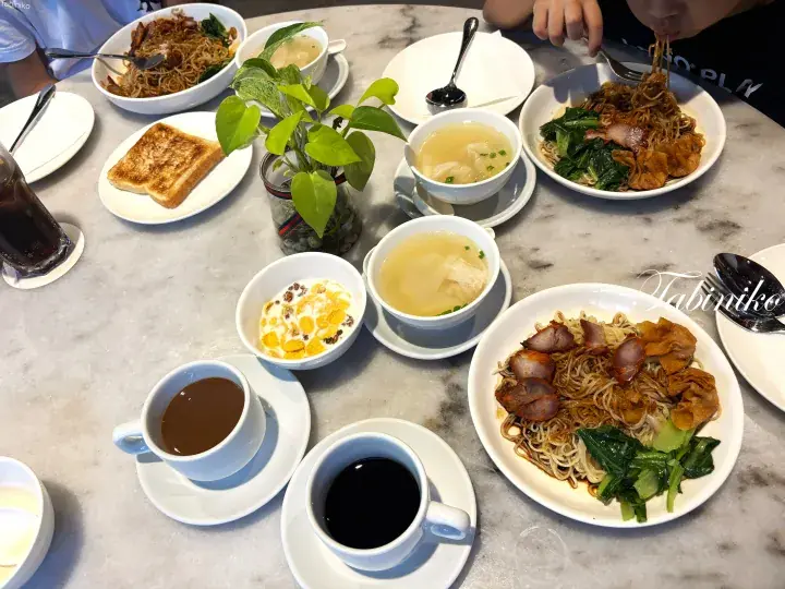A table spread with Malaysian dishes including noodles, toast, soup, and drinks.