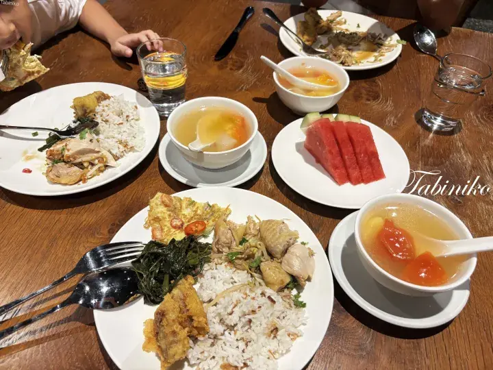 A wooden table set with plates of Hainanese chicken rice, soup, watermelon, and a child eating.