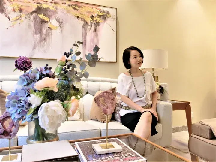 A woman sits on a white sofa in a modern living room with abstract art and a large floral arrangement.
