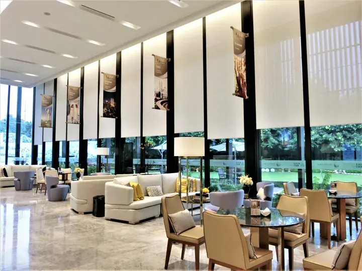 Bright hotel lobby with white sofas, beige chairs, and floor-to-ceiling windows overlooking a lush green garden.