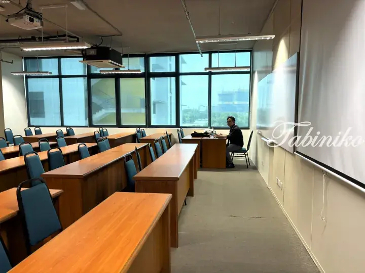 Modern lecture hall with wooden desks and blue chairs, large windows, and a person sitting at a desk near a whiteboard.