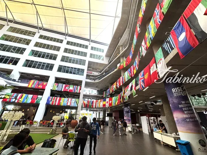Students walking through a modern atrium decorated with numerous international flags at a Malaysian university campus.