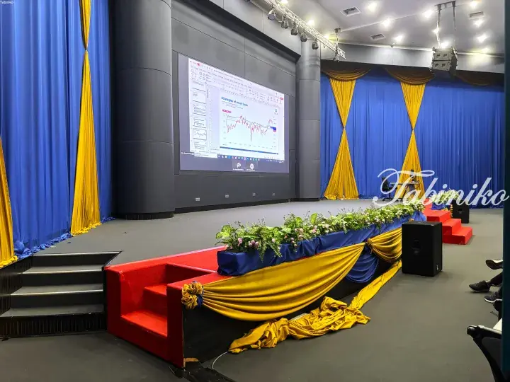 Stage decorated with blue and yellow drapes and floral arrangements for an event, with a presentation screen visible.