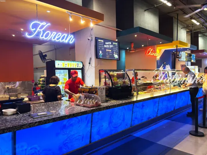 Korean and Chinese food stalls with neon signs at a modern Malaysian food court counter.