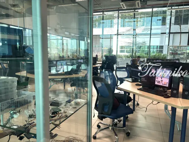 Modern open-plan office with glass walls, computer workstations, and blue ergonomic chairs.