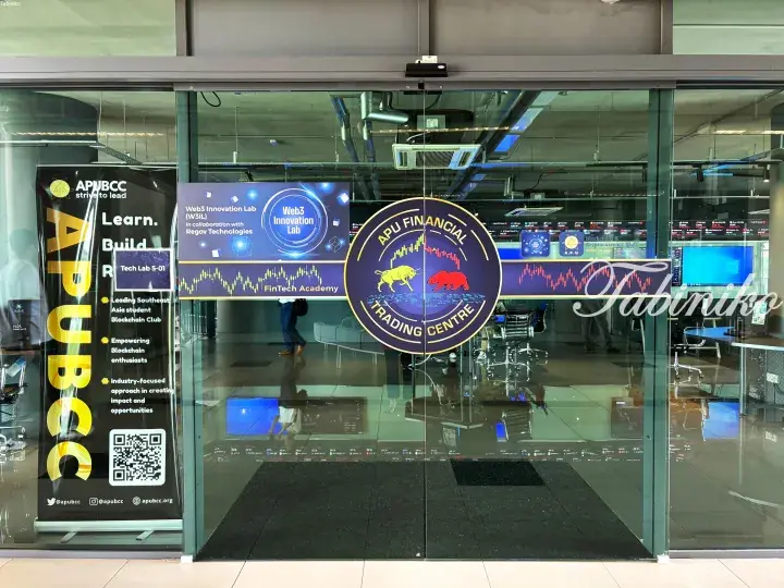 Glass entrance to APU Financial Trading Centre with bull and bear logos, plus a banner for APU BCC and Web3 Innovation Lab.