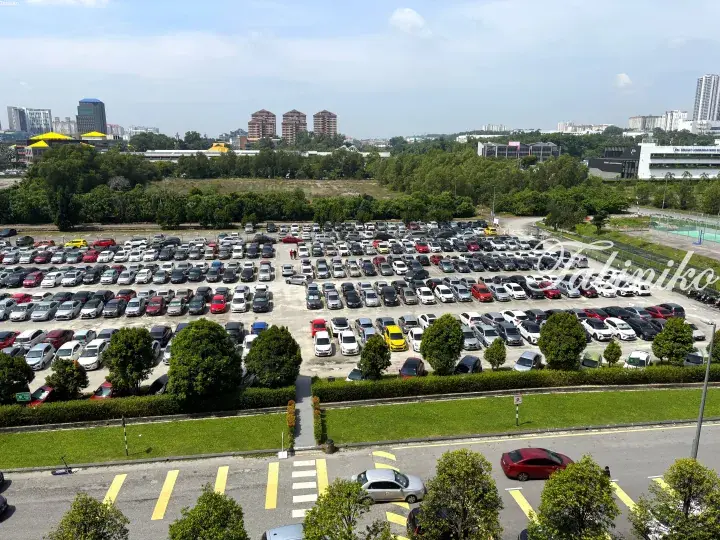 Aerial view of a large parking lot filled with cars, surrounded by greenery and urban buildings in the background.