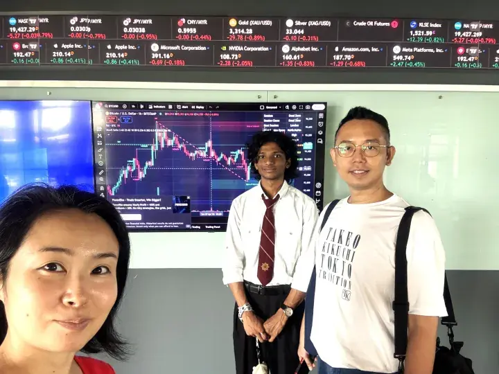 Three people pose in front of a large screen displaying financial stock market charts and data.