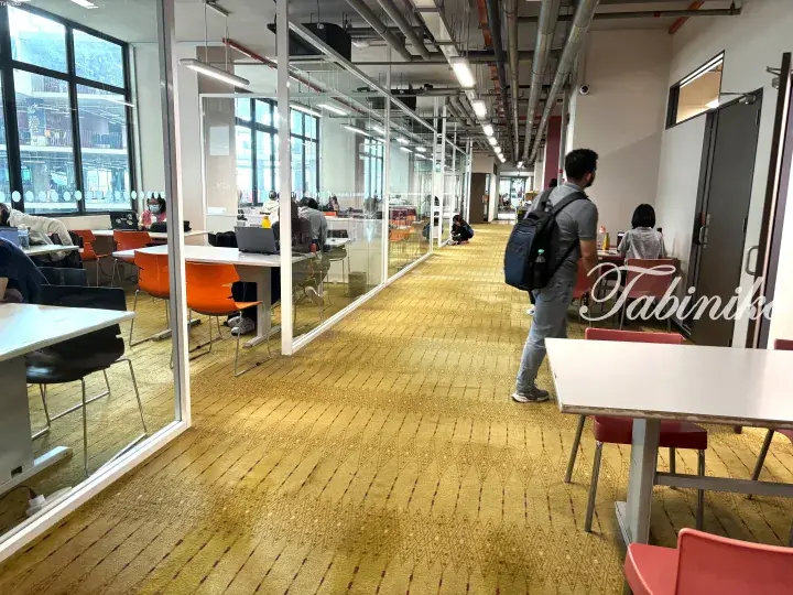 Modern coworking space with glass-walled meeting rooms, orange chairs, and a person walking through the hallway.