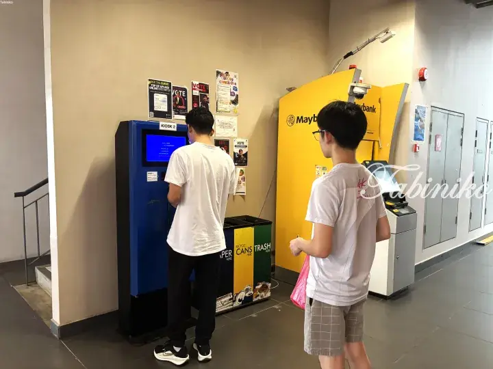Two young men using a blue self-service kiosk and a yellow Maybank ATM inside a modern building lobby.