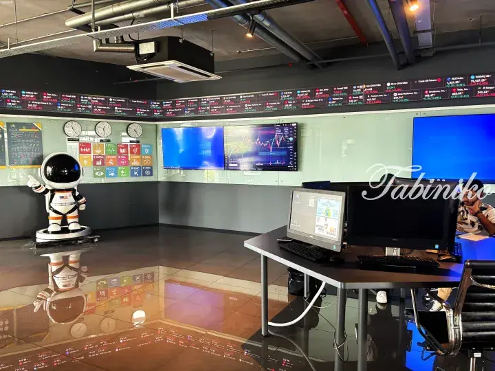 Modern office space with stock market ticker displays, dual monitors, and a decorative astronaut figure.