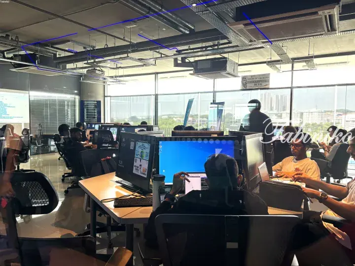 Modern open-plan office with employees working at desks near large windows overlooking a cityscape.