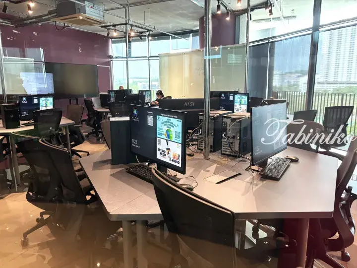 Modern co-working space with rows of desktop computers and ergonomic chairs near large windows.