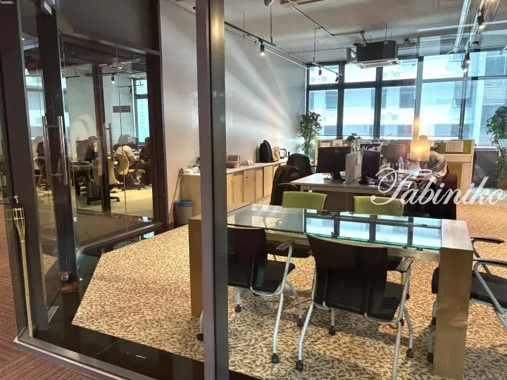 Modern office interior with glass meeting table, chairs, and workstations near large windows.