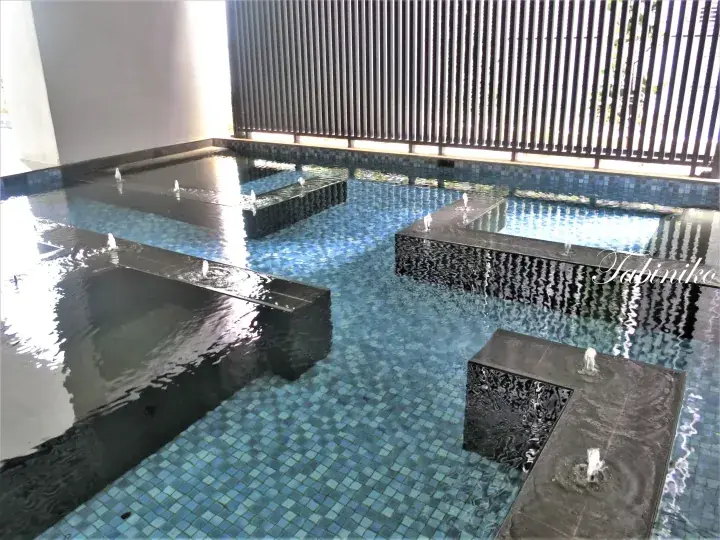 Modern indoor pool with blue mosaic tiles and black marble platforms featuring small water fountains.
