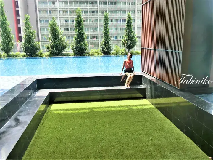 Woman relaxing on a dark tiled ledge beside a rooftop infinity pool with artificial grass and city views.