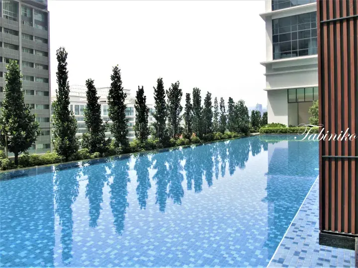 Modern infinity pool with blue tiles reflecting tall trees and high-rise buildings at a luxury condominium.