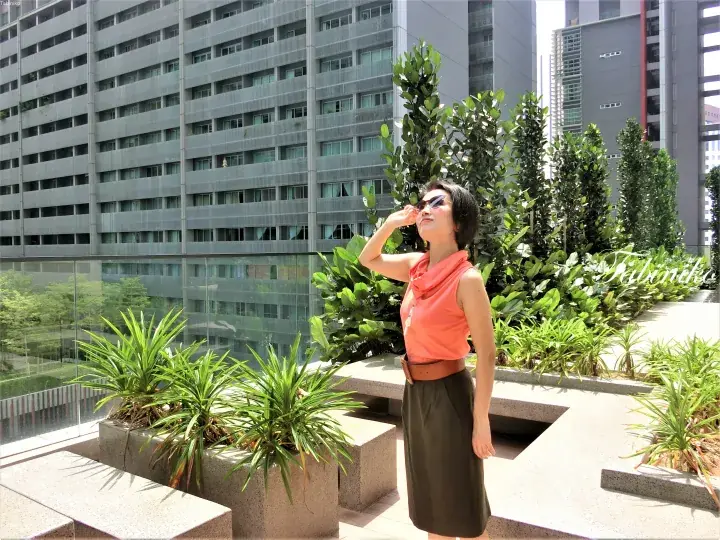 Woman in orange top and brown skirt shielding her eyes on a sunny rooftop garden with modern high-rise buildings in the background.
