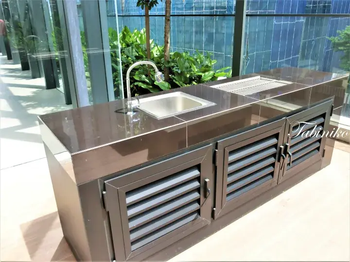 Modern outdoor kitchen island with dark brown cabinets, stainless steel sink, and louvered doors in a tropical setting.