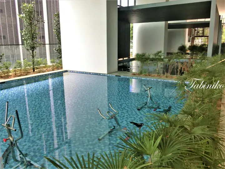 Indoor swimming pool with stationary bikes submerged in the water, surrounded by tropical plants and modern architecture.