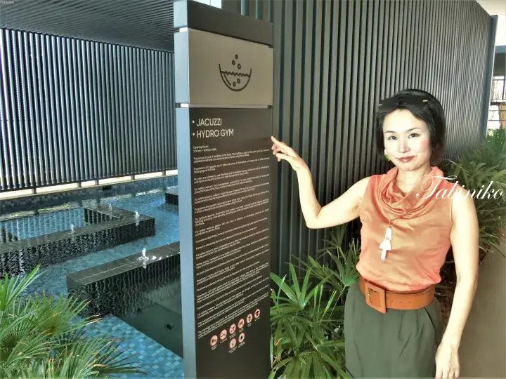 Woman pointing to a sign listing jacuzzi and hydro gym amenities at a modern poolside facility.