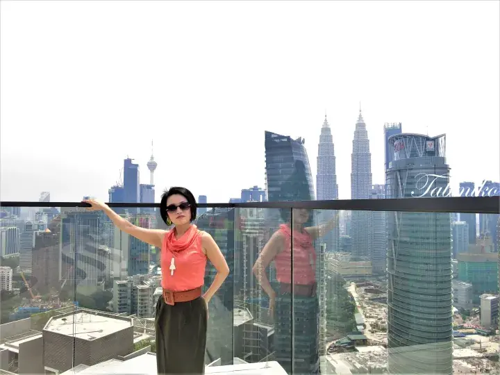 Woman in sunglasses posing on a balcony with the Kuala Lumpur skyline and Petronas Towers in the background.