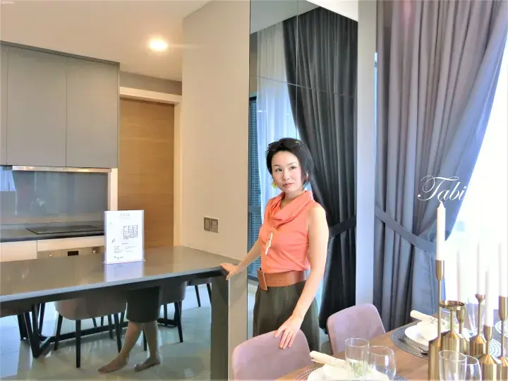 Woman standing in a modern kitchen and dining area with sleek grey cabinetry and large windows.