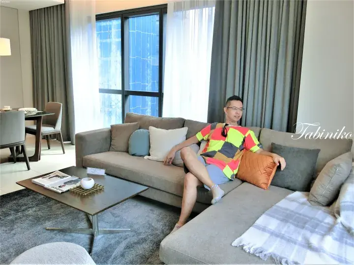 Man relaxing on a modern grey sectional sofa in a spacious, well-lit living room with floor-to-ceiling windows.