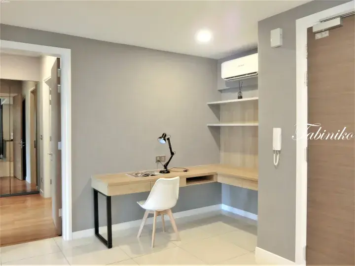 Modern L-shaped study desk with white chair, lamp, and shelves in a minimalist apartment room.