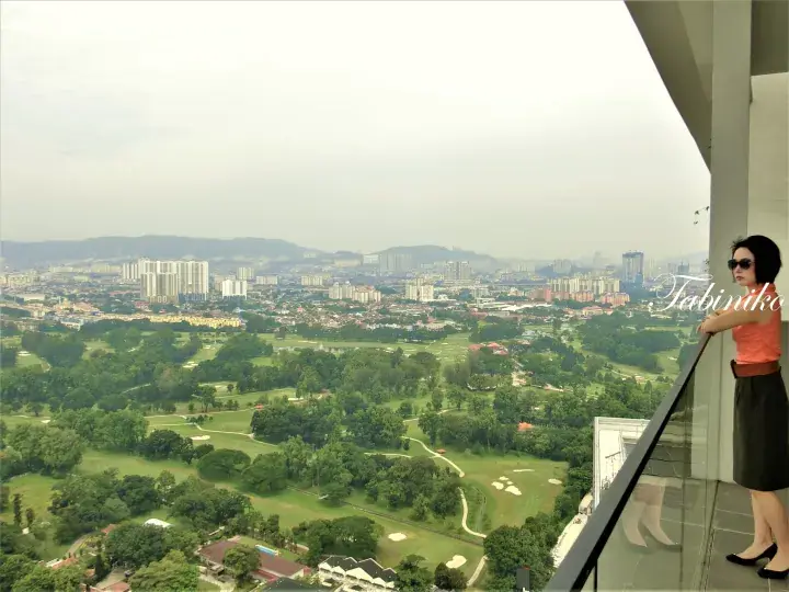 Woman on a high-rise balcony overlooking a lush golf course and city skyline in Malaysia.