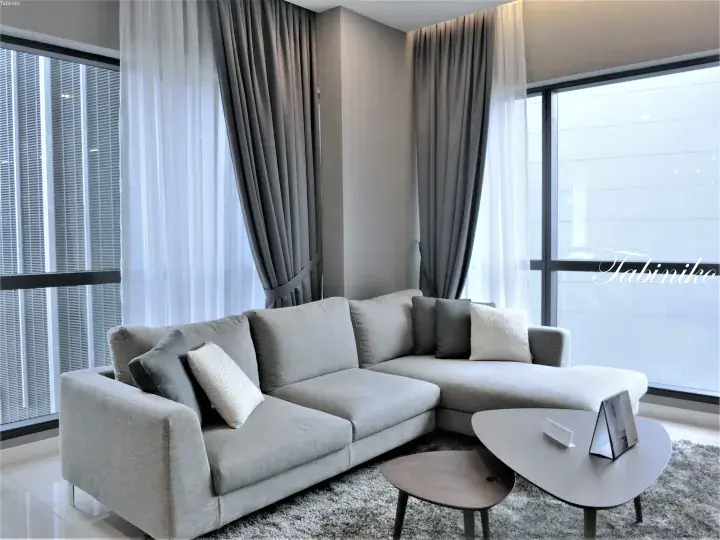 Modern L-shaped sofa in a bright living room with floor-to-ceiling windows and grey curtains.