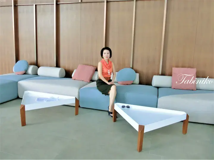 Woman in orange top and green skirt sitting on a modern blue and white sectional sofa in a minimalist lounge area.