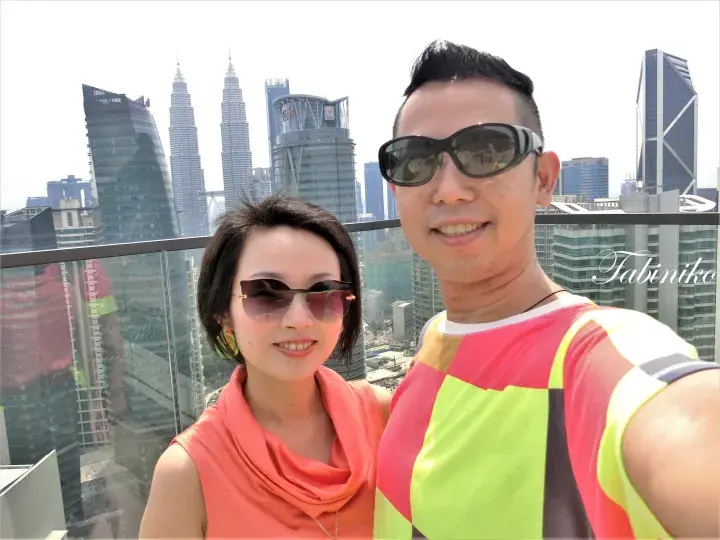 Couple taking a selfie on a balcony with the iconic Petronas Twin Towers and Kuala Lumpur skyline in the background.