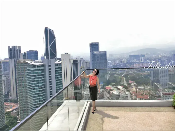 Woman in orange top and black skirt posing on a high-rise balcony with a panoramic view of Kuala Lumpur’s city skyline.