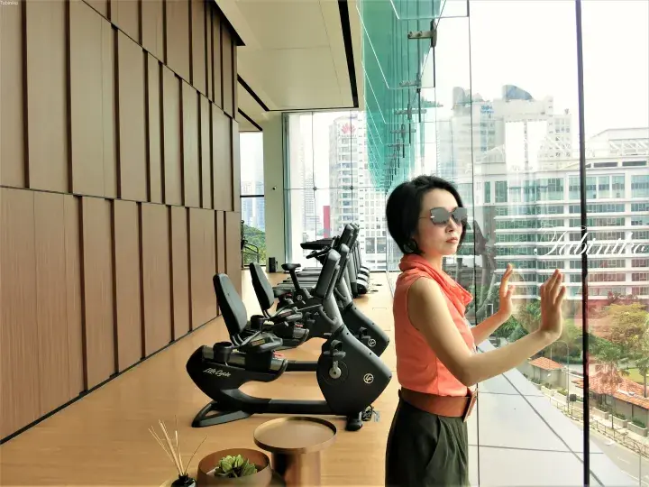 Woman in sunglasses and orange top standing by a window with a city view inside a modern gym with exercise bikes.