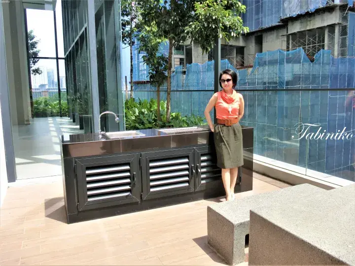 Woman in orange top and green skirt standing beside a modern outdoor kitchen counter with a sink and grill.