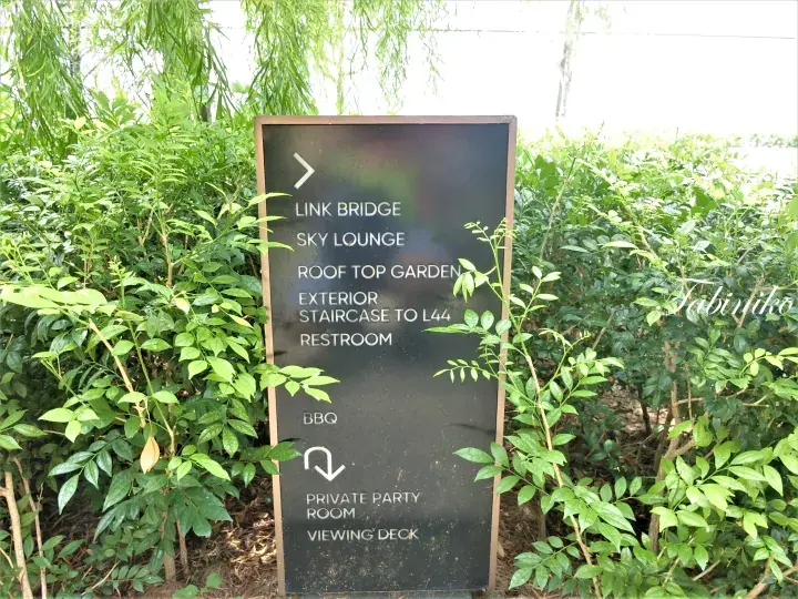 Black directional sign listing amenities like Sky Lounge and Rooftop Garden amidst lush greenery.