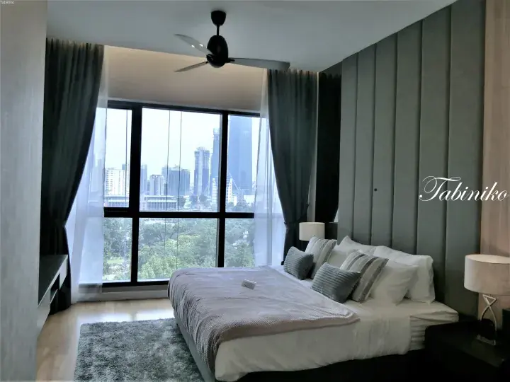 Modern bedroom with city skyline view from KLCC area, featuring a large bed, grey wall paneling, and ceiling fan.