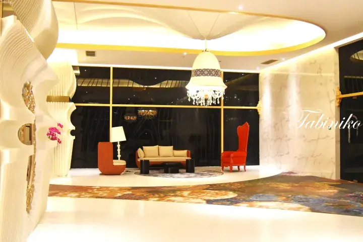 Luxury hotel lobby featuring a white sculptural wall, red high-back chair, beige sofa, and elegant chandelier.