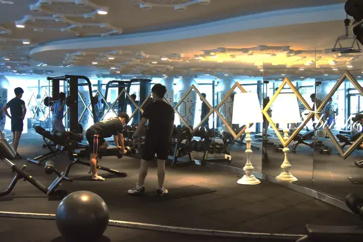 People exercising with weights in a modern gym with mirrors and decorative ceiling lights.