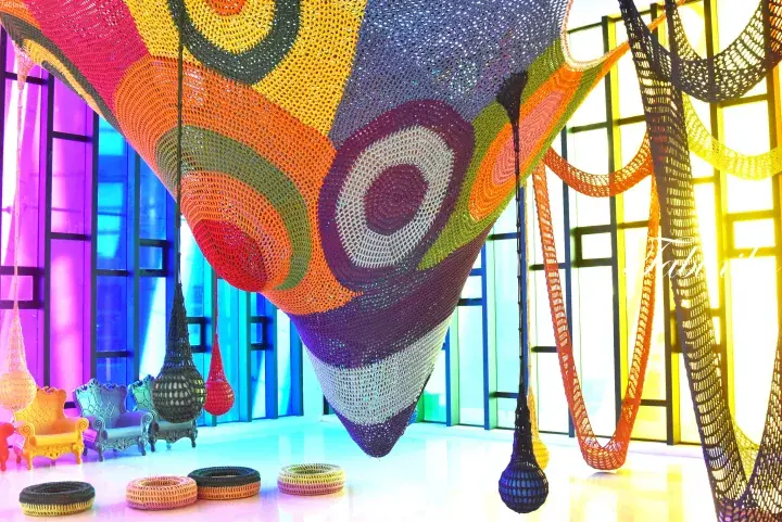 Colorful woven hanging installations and chairs in a modern interior space with large windows.