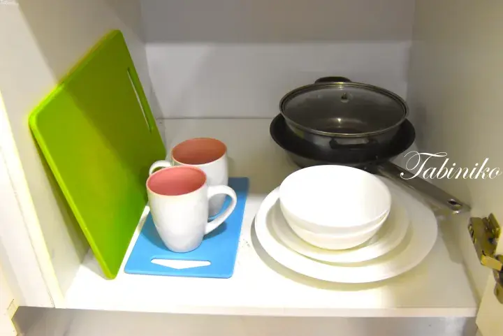 Kitchen cabinet with green cutting board, blue mat, two mugs, stacked white bowls, and a pot with lid.