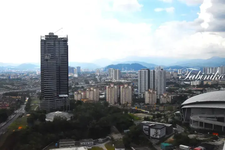 Kuala Lumpur skyline featuring a tall dark skyscraper, residential towers, and a stadium against a backdrop of mountains.