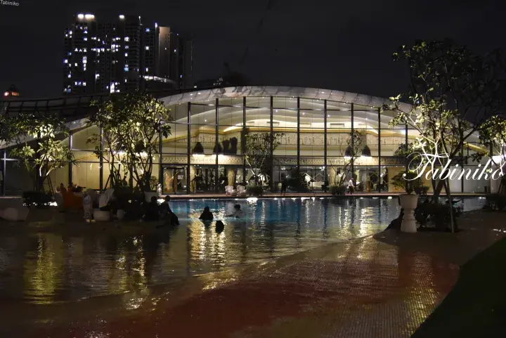 Night view of a modern glass building with a swimming pool and city skyline in the background.